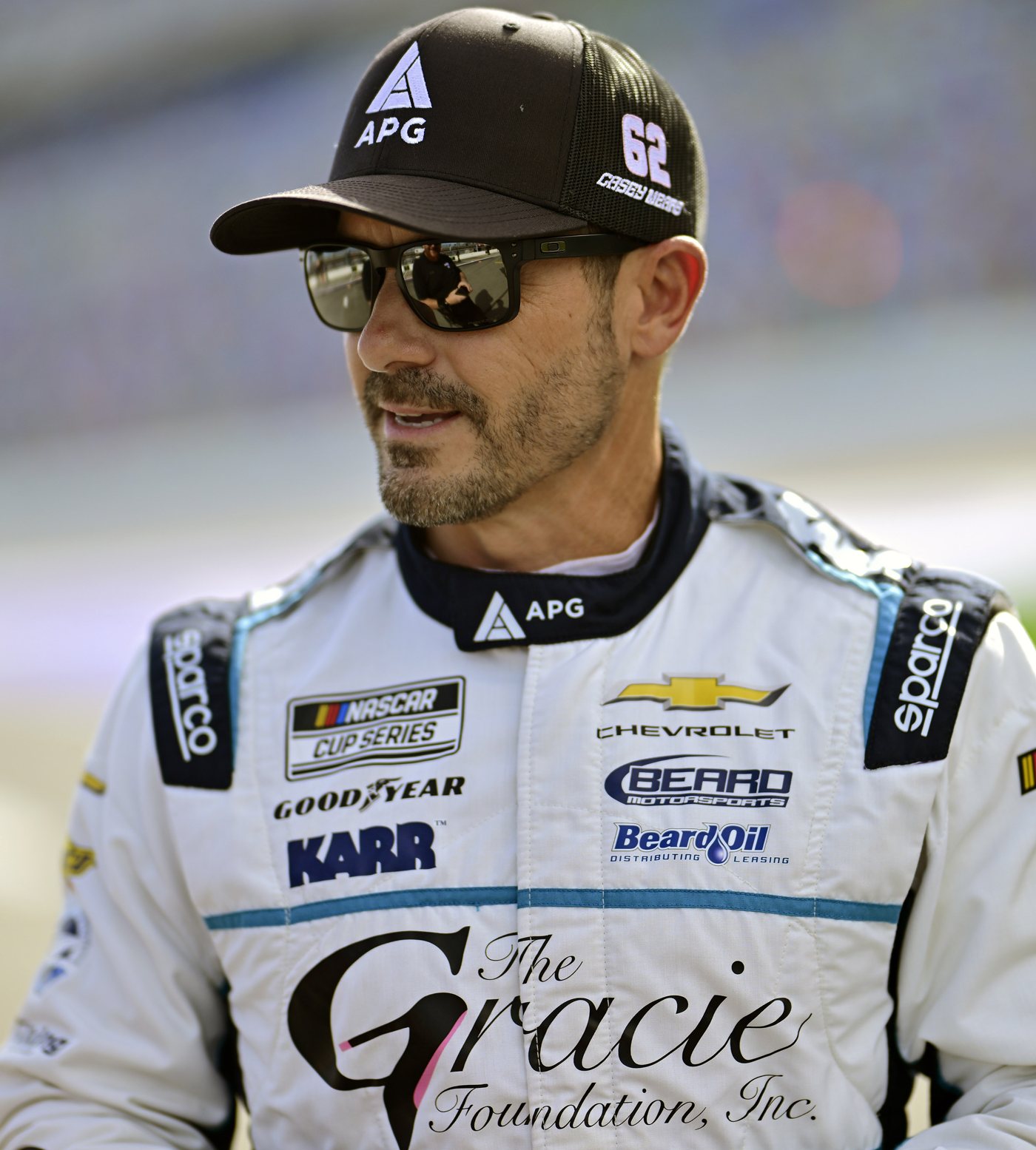 Casey Mears in his white firesuit with The Gracie Foundation and APG logos, wearing an APG cap and sunglasses