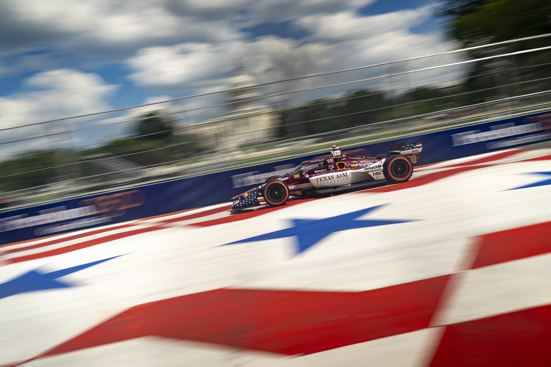 The maroon Texas A&M No. 60 IndyCar at speed on a street circuit painted with stars and stripes, the U.S. Capitol blurred behind