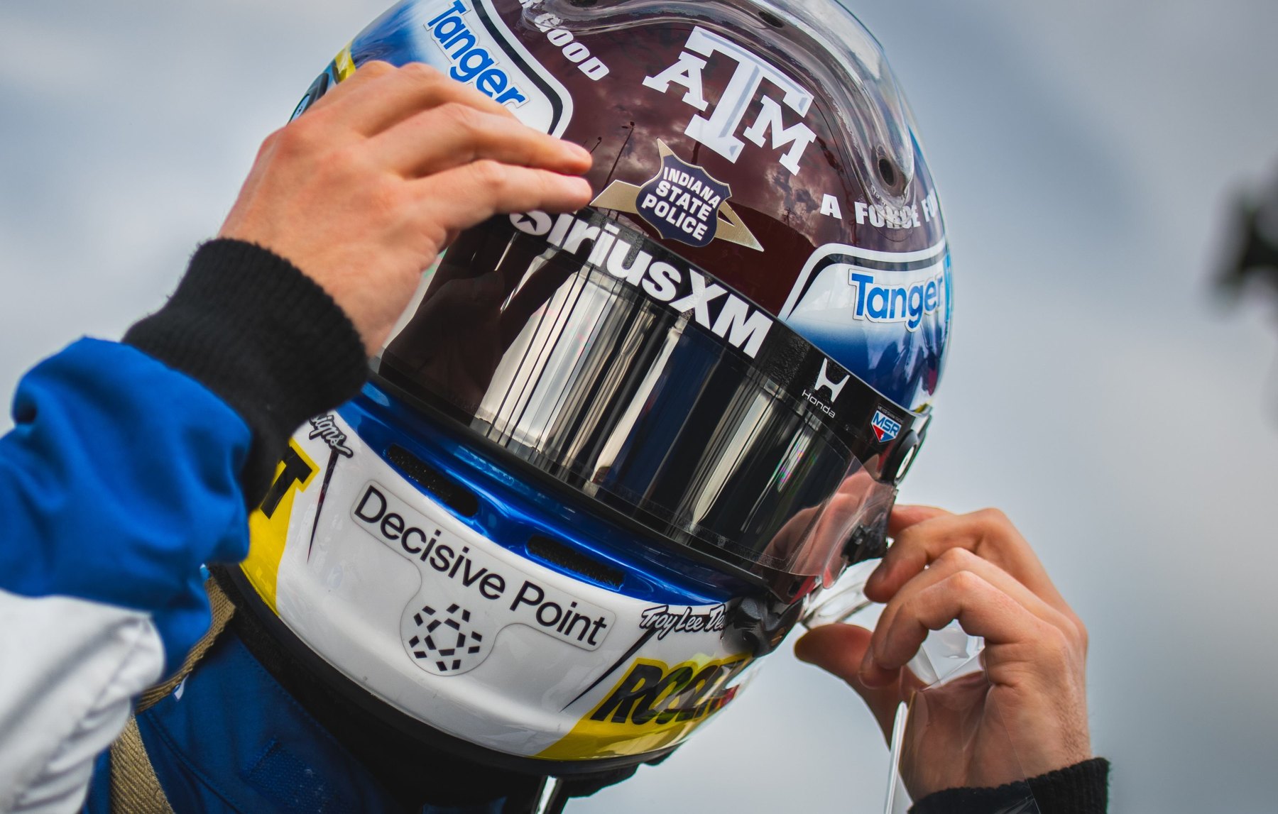 Felix Rosenqvist fastening his helmet, which carries the Texas A&M, Indiana State Police, Decisive Point and Tanger logos